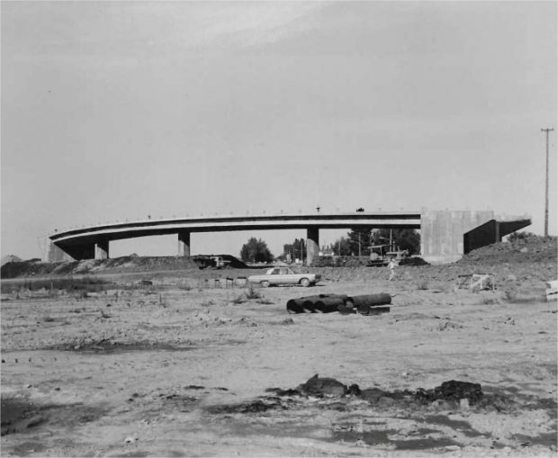 A highway under construction with houses and vehicles circulating in the background