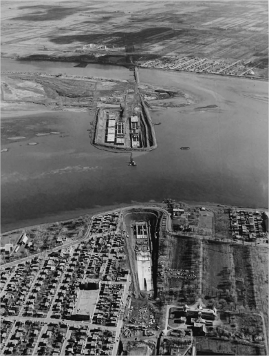 A construction site in the middle of a river with a city and fields in the background