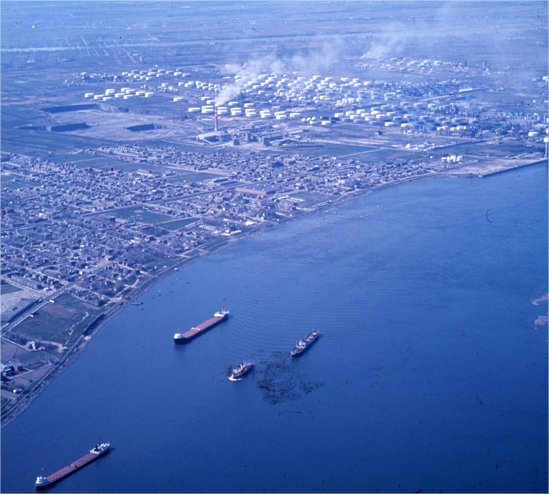 Boats sailing on a river near a city with oil refineries in the background