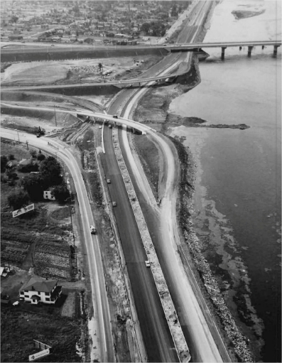 A highway under construction with houses and vehicles circulating in the background