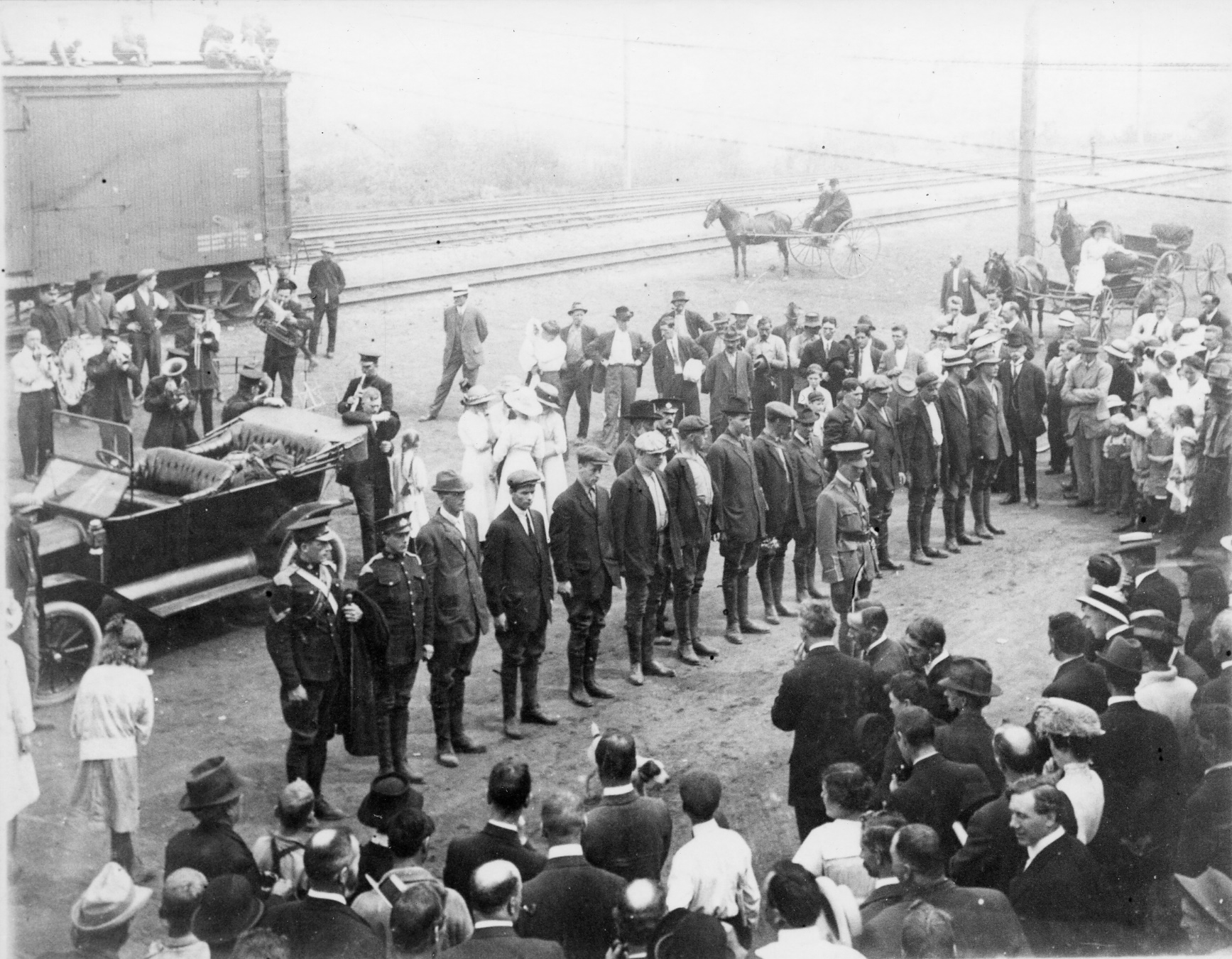 A line of men and soldiers pose facing a crowd in front of train tracks