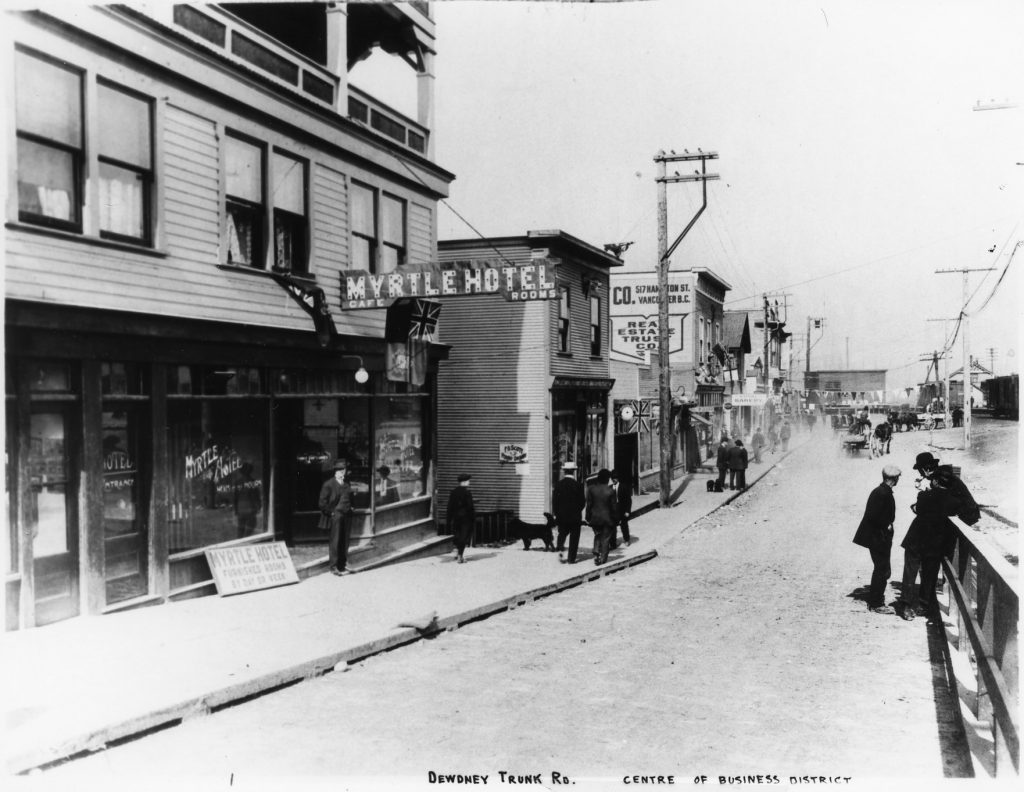 A black and white city street with buildings on one side and people walking on the sidewalk beside them.