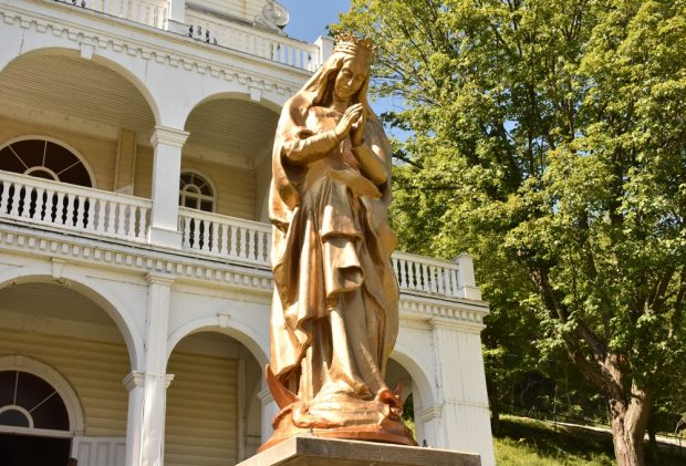 Colour photo of a large, golden-coloured statue of the Virgin Mary wearing a crown and standing on a dragon, her hands clasped in prayer. Behind the pedestal-mounted statue, a multi-storey building can be seen.