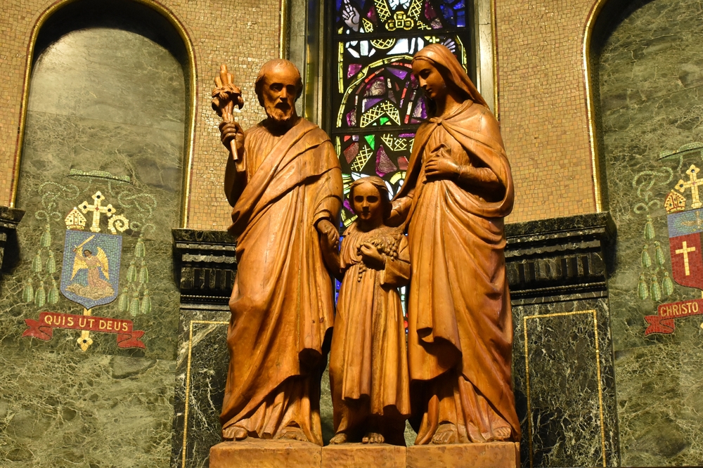 Colour photo of a large wooden sculpture depicting (from left to right) Joseph, Jesus, and Mary. The sculpture is mounted on a pedestal in a dark, ornately decorated chapel.