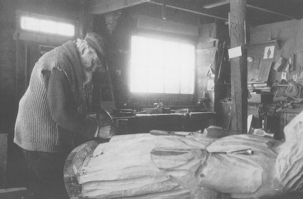 Black and white archival photo. A man sculpts the foot of a large wooden statue in a workshop where drawings and woodworking tools can be seen.