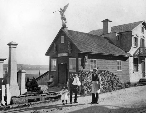 Black and white archival photo. Two men are seen standing in front of a building consisting of two sections. A sign above the door on the single-storey part on the left reads “L. JOBIN.” The two-storey section on the right is the home. The man on the left is accompanied by two young children.