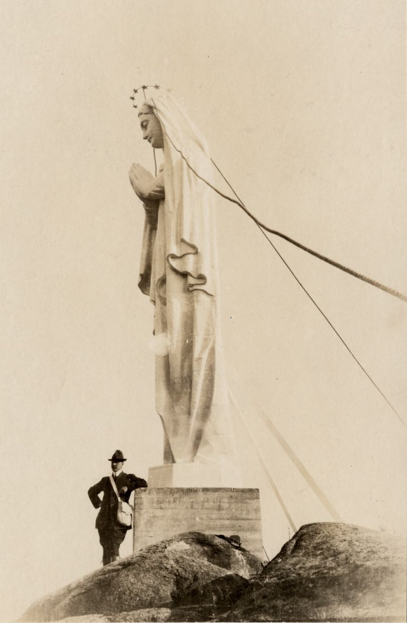 Black and white archival photo. A man leans against the foot of a statue that stands several times his height. The white sculpture of the Virgin Mary has a crown of stars, and her hands are clasped in prayer. The statue is mounted on a pedestal built directly into the rock.
