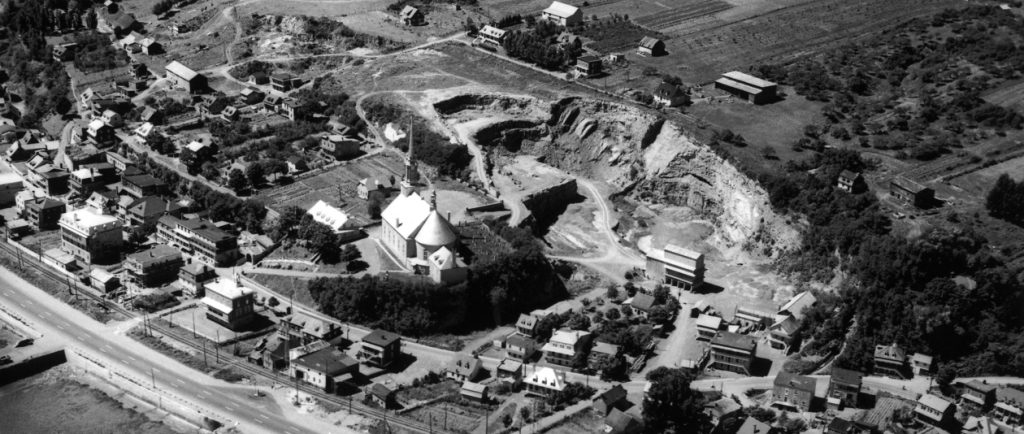 Black and white archival photo. The municipality of Château-Richer radiates around the church, seen in the centre of the picture. In addition to houses, we can see a stone quarry, cultivated fields, a railroad track, and Boulevard Sainte-Anne.