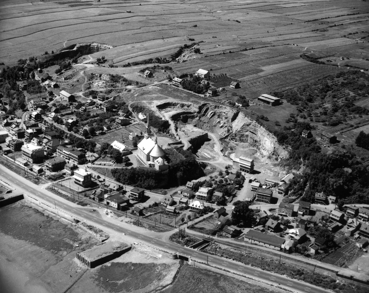 Black and white archival photo. The municipality of Château-Richer radiates around the church, seen in the centre of the picture. In addition to houses, we can see a stone quarry, cultivated fields, a railroad track, and Boulevard Sainte-Anne.