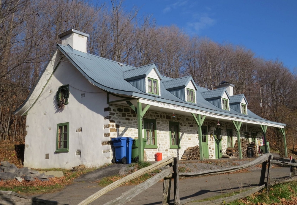 Colour photograph of a two-storey heritage home built directly on the ground. The home has two chimneys, one at either end of its gabled roof that features four dormer windows at the front. The stone walls are covered for the most part with pale-coloured plaster, while the façade has more exposed stones. There are five windows and two doors on the main floor, painted a pale green colour.