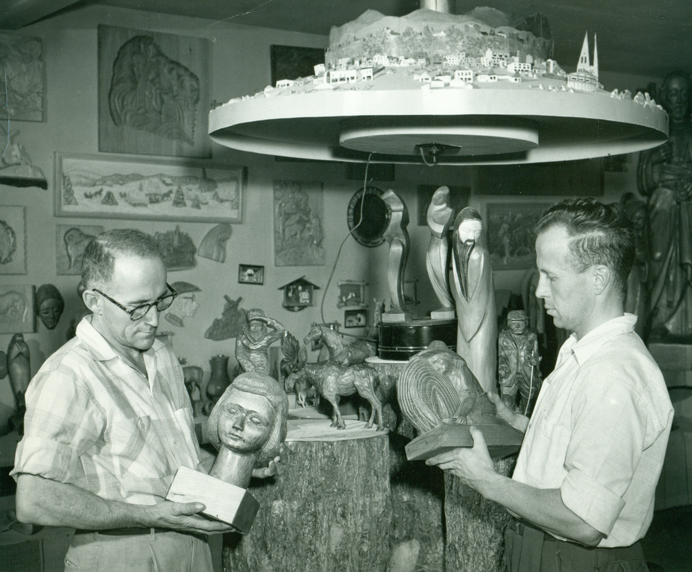 Black and white archival photo showing Alphonse Paré (left) and Louis-Philippe Paré (right) holding wooden sculptures. Some of the artist’s works can also be seen on the walls and surfaces around the two men.