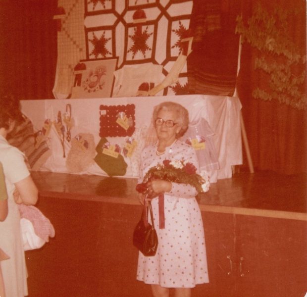 Colour photo of Amanda Gagnon Jalbert holding a bouquet of flowers. She is wearing a white print dress and holding a handbag. On the stage behind her, textile handicrafts are displayed on a table.
