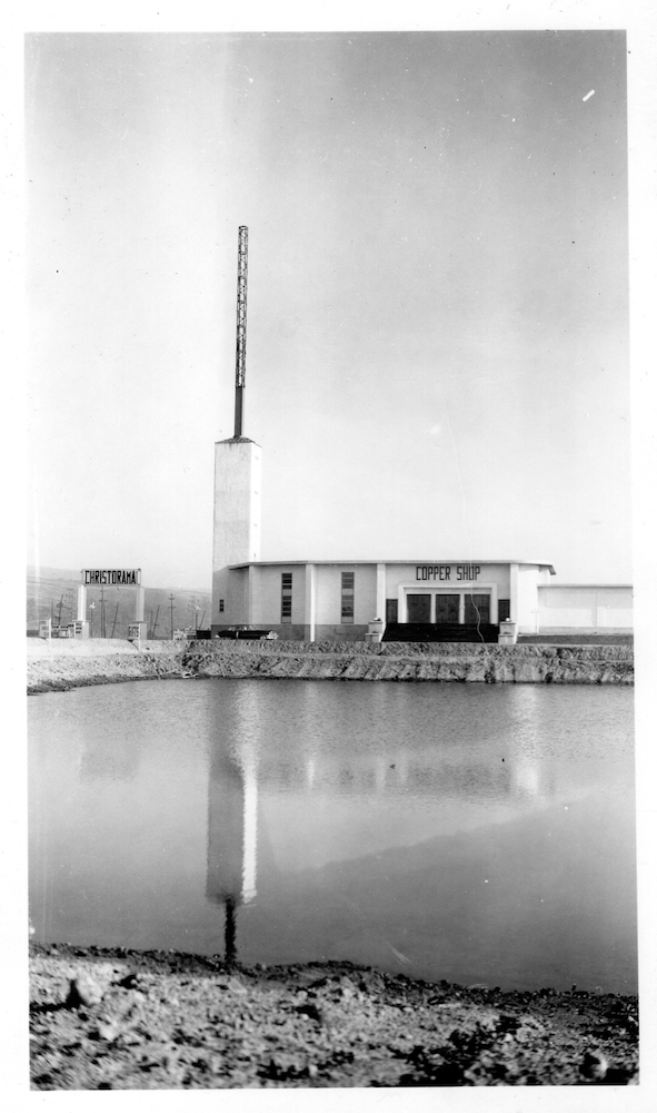 Black and white archival photo showing a modern, two-storey building beside a road, with a chimney at the far left. In front, the building can be seen reflected in a body of water. On the left, a large sign reads “Christorama.” It also says “Copper Shop” and “atelier de cuivre” on the building.