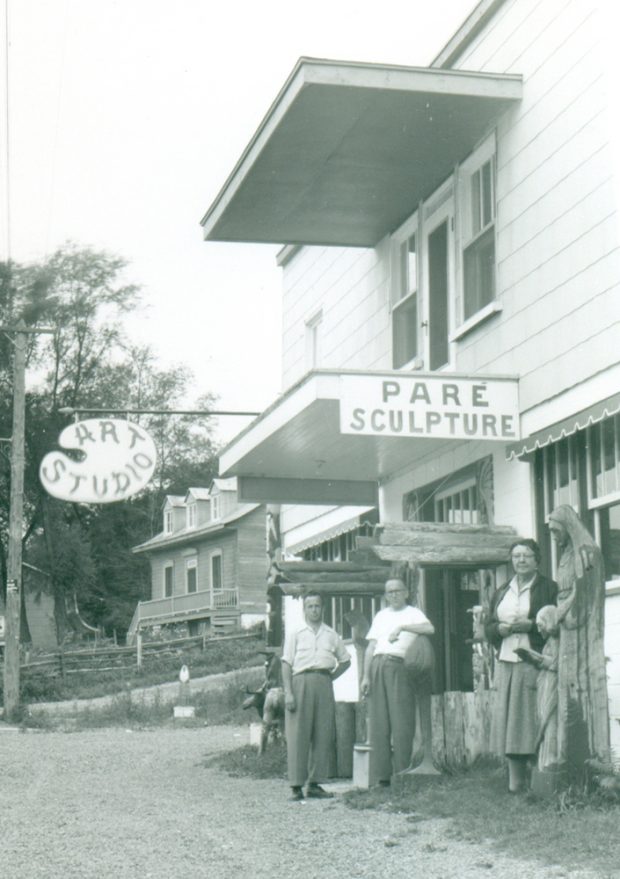 Black and white archival photo. Louis-Philippe Paré, Alphonse Paré, and an unidentified woman are standing facing the camera by the side of the road in front of a two-storey building. Two signs on the façade of the building read “Paré Sculpture” and “Art Studio.” A house can be seen in the background. Several wooden sculptures stand in front of the building, on either side of the front entrance.