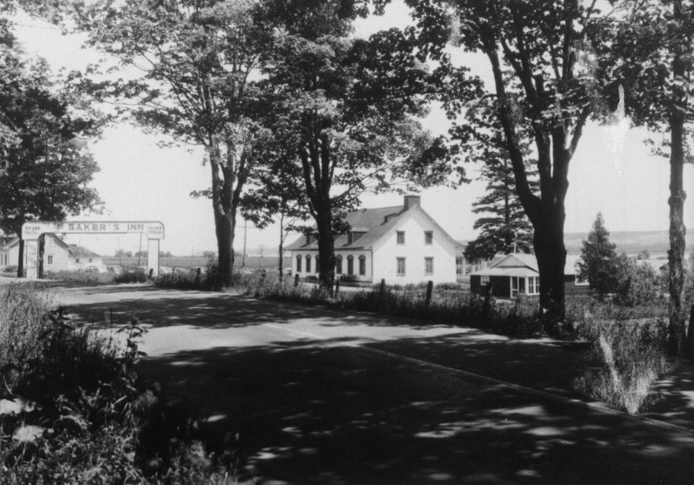 Black and white archival photo showing a signpost arch over the lane leading to a white inn that can be sign from a side angle on the right. It reads “The Baker’s Inn.” The inn has a gabled roof with three dormers in front and a chimney at either end. There are six windows on the front of the building. Mature trees line the lane leading to the inn.