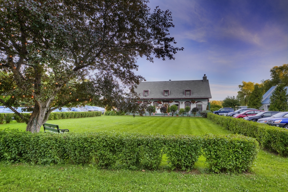 Colour photo showing Auberge Baker in the background, with its three dormers and six windows on the front. In the foreground, the inn’s grassy lawn is bordered by a row of shrubs and a mature tree. Part of the parking lot and several vehicles can be seen on the right of the photo.