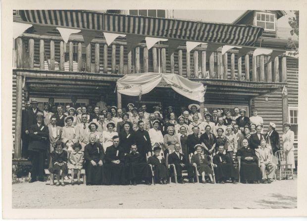Black and white archival photo of a large group gathered in front of Auberge du Lac. The guests are seated or standing in five lines, and they include members of the clergy, young children, women, and men. The log building behind them stands two storeys tall. The upper floor has a wooden balcony from which a decorative banner hangs.