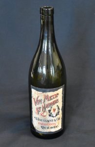 Colour photo of a long-necked wine bottle against a black background. The bottle bears a label that translates as “St. Nazaire Sacramental Wine, A. Toussaint & Cie Manufacturers, Québec.”