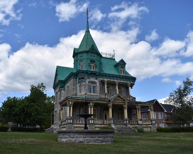 Colour photo of an imposing, three-storey stone home set in the middle of a grassy lawn. The roof is green. The façade of the house features an upper balcony and a veranda on the ground floor. The window frames, posts, and balustrade are painted yellow. There is a fountain in front of the house, and in the background, we see trees and a neighbouring house.