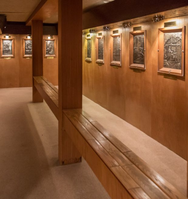 Colour photo showing a wood-panelled room featuring a series of tableaus in embossed silver. Above each tableau is a short description. In the middle of the room, a long bench seat intersected by a column stretches from one side to the other.