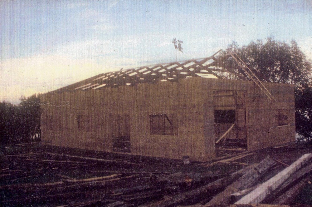 Colour archival photo of a building under construction. We can see the wooden walls as well as the roof trusses, also in wood. In the foreground and alongside the building are piles of lumber and logs.