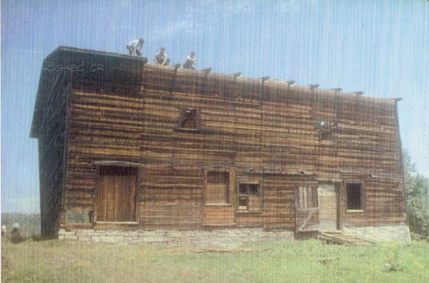 Colour archival photo of a wooden barn with stone foundations. We can see two windows on the upper floor and two doors and three windows on the lower floor. Three men are at work taking the roof apart.