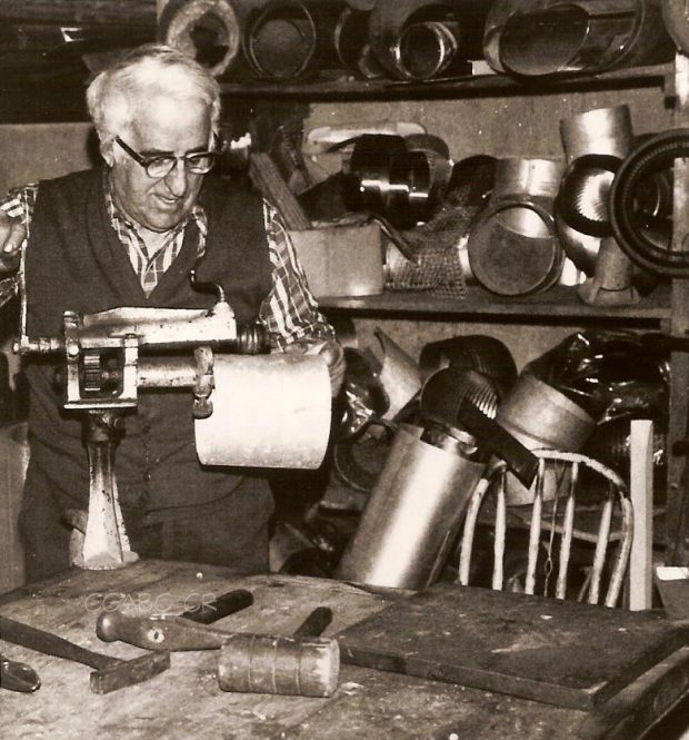 Black and white archival photo depicting Joseph Drouin at work in his workshop. He is operating a machine with his right hand while holding the object he is making in his left. On the table in front of him are a number of tools such as hammers and a mallet. The shelves in the background are piled high with various metal objects.