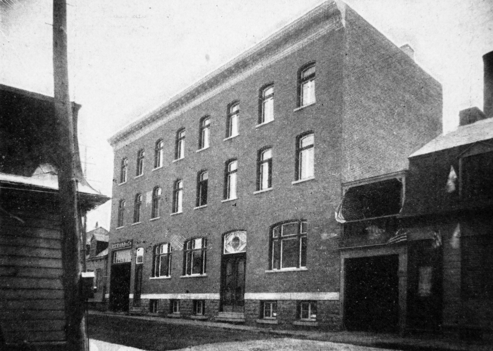 Photographie d’archives en noir et blanc montrant un édifice de trois étages qui inclut un sous-sol. Les deux étages supérieurs sont percés de 8 fenêtres chacun. Au rez-de-chaussée, la façade compte trois fenêtres et trois portes, dont une plus importante à gauche. Au-dessus de celle-ci, une enseigne indique « Richard & Cie ». L’édifice a des murs mitoyens avec les édifices voisins.