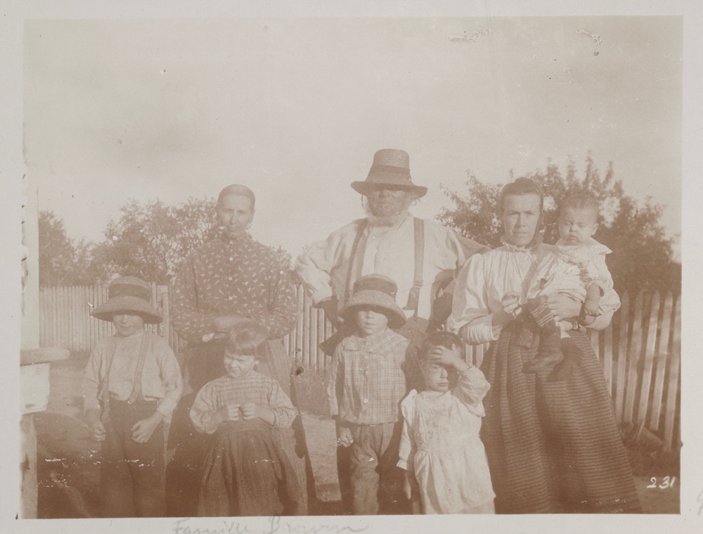 Black and white archival photo of a family standing outside, with a fence and some trees in the background. Hugh Brown is in the centre of the photo wearing a hat. A woman stands on either side of him. The one on the right is holding an infant in her arms. Four young children are standing in front of the adults.