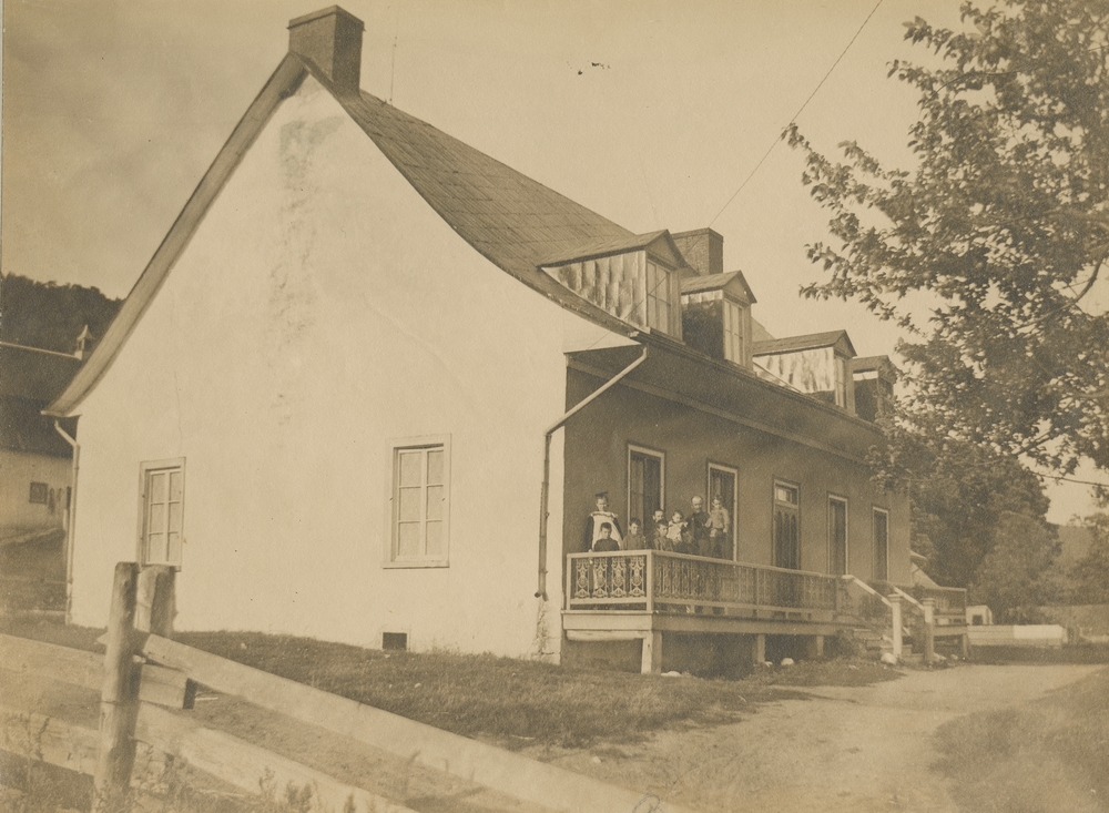 Black and white archival photo (side view) of a white house with a gabled roof and a front balcony. We can see two chimneys and four dormer windows. On the façade there are two windows on either side of the front door. A family of seven children poses for the camera on the balcony. In the foreground there is a tree and a lane in front of the house.