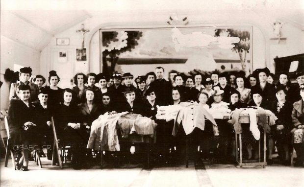 Black and white archival photo showing a group of two rows of women posing for the camera in a large room. Those in the front row are seated, with the others standing behind. The only man in the photo, a priest, stands in the centre. Various textile handicrafts are displayed on tables in front of the group.