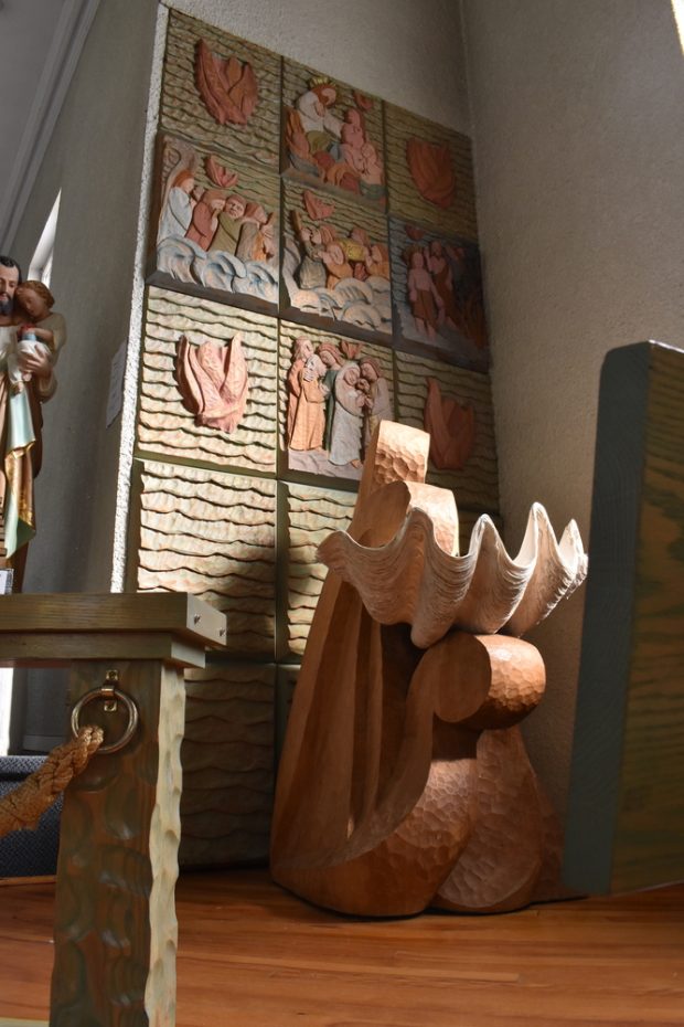 Colour photo of the inside of a church. The baptismal font in the centre of the photo consists of a huge seashell with a base carved in wave-like patterns. Behind it on the wall, several sculpted and painted tableaus depict scenes from the Bible in which water and waves are a key motif.