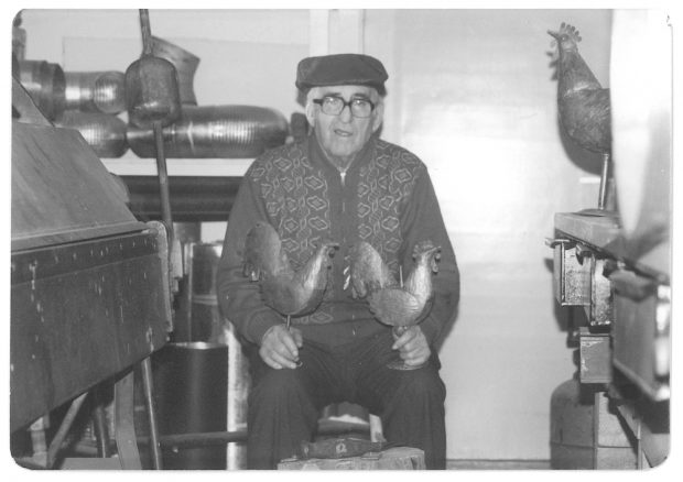 Black and white archival photo of Joseph Drouin in his later years, seated in his workshop and holding two cooper roosters on his lap. On a shelf behind him on the left, we can see pieces of metal pipe. On the right, a larger copper rooster perches on his workbench.