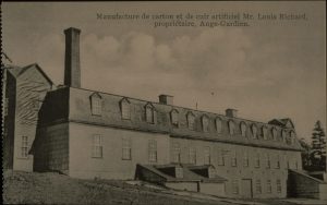 Photographie d’archives en noir et blanc montrant une vue de trois-quarts d’un bâtiment industriel à trois étages percés de multiples fenêtres et à toit mansardé. Une longue cheminée se dresse à l’arrière gauche du bâtiment. Deux cheminées moins hautes s’élèvent au centre de l’édifice.