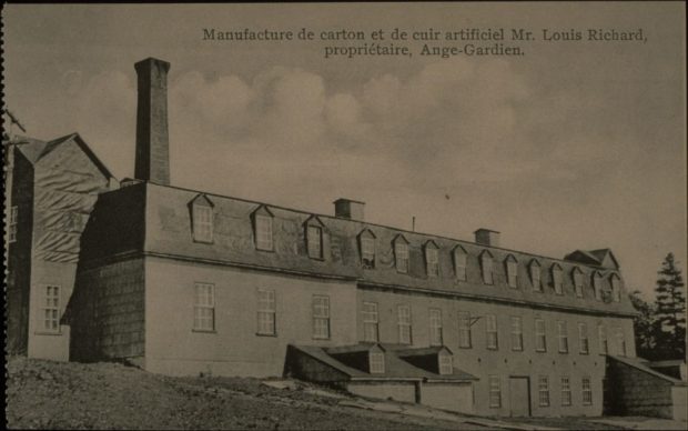 Black and white archival photo showing a three-quarter view of an industrial, three-floor building with multiple windows and a mansard roof. A tall chimney stands behind the building to the left. Two lower chimneys emerge from the centre of the factory.
