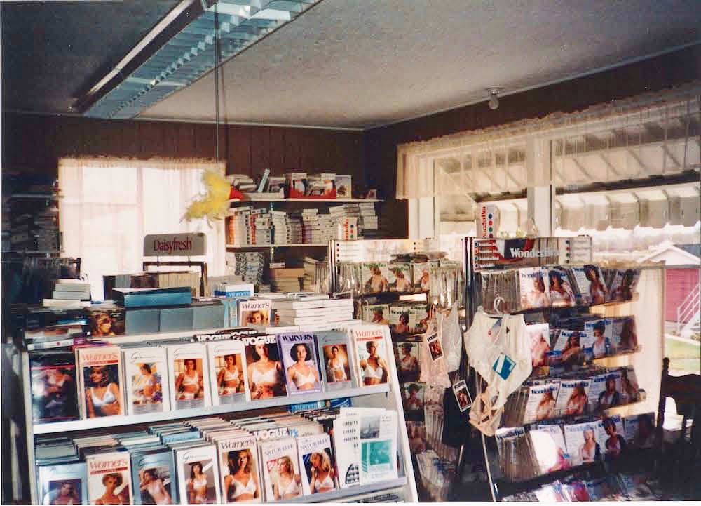 Colour photo showing racks of goods for sale. We can see boxed bras and other undergarments on display racks and on shelves along the walls. Two windows allow in daylight.