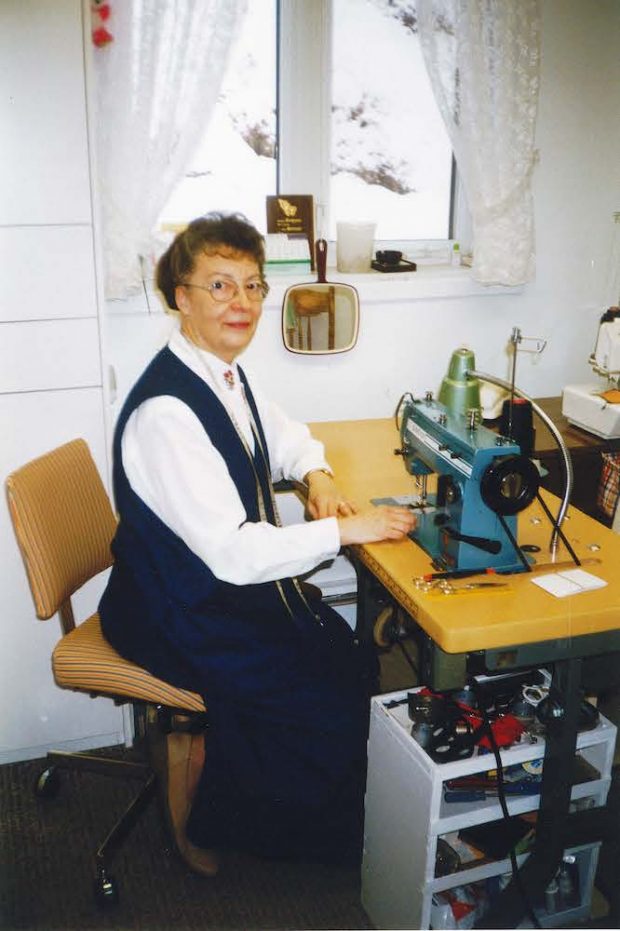 Colour photo of Marie-Rose Jalbert sitting by a window at her sewing machine. She has a measuring tape hanging around her neck, and we can see sewing accessories, a mirror, and a lamp on her worktable.