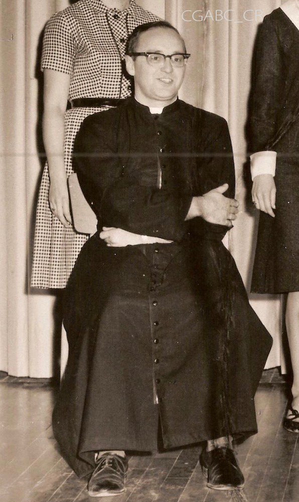 Black and white archival photo showing vicar Jean Moisan sitting with his arms crossed. He is wearing glasses, a cassock, and dark-colour shoes. Two women, their faces partially hidden, are standing behind him.