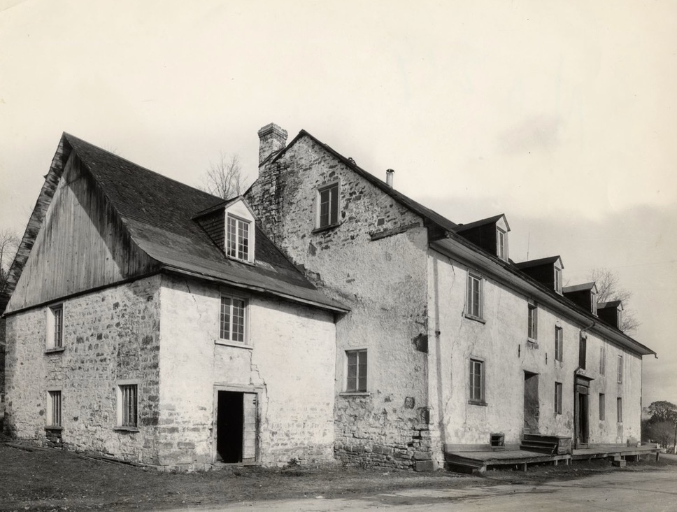 Black and white archival photo showing a distant view of the façade and left profile of a three-storey gabled building made of stone partially covered with lime mortar. A smaller annex can be seen on the left.