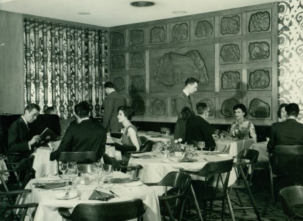 Black and white archival photo depicting a dining room with patrons sitting at tables and waiters serving them. On the back wall is a sculpted wood mural consisting of a central panel surrounded by twenty-two smaller panels.