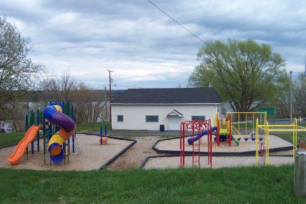 Photo of a park showing a small white building with a door and three windows on the façade looking over a series of playground modules. On the left is a spiral slide and a straight slide, as well as climbing modules, while on the right we can see swings and a smaller slide. The park is surrounded by grass.