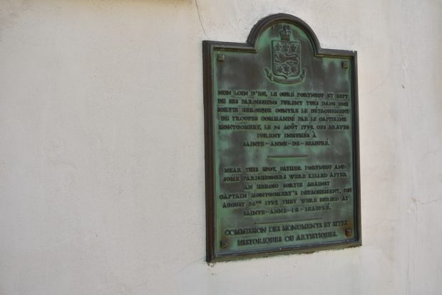 Colour photo of a bronze plaque with the French inscription followed by an English translation reading “Near this spot, Father Portneuf and some parishioners were killed after an heroic sortie against Captain Montgomery’s détachement, on August 24th, 1759. They were buried at Sainte-Anne-de-Beaupré.” At the bottom of the plaque, it reads: Commission des monuments et sites historiques ou artistiques.