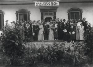 Photographie d’archives en noir et blanc montrant un couple de mariés entourés de leurs élégants invités devant l’auberge Baker. Tous regardent la caméra. Derrière le groupe, au-dessus de la porte, on voit une enseigne qui indique « ALL YOU CAN EAT 0,50$ ». L’avant-plan de la photographie est occupé par quelques plantes et arbustes.