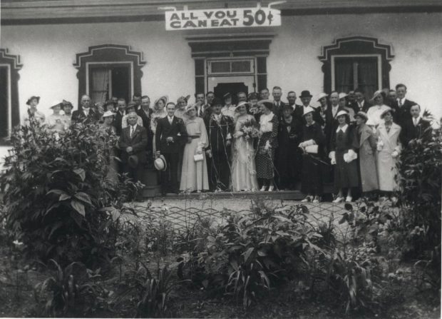 Black and white archival photo showing a bride and groom surrounded by their elegantly dressed guests in front of Auberge Baker. Everyone is looking at the camera. Behind the group, above the door, hangs a sign reading “ALL YOU CAN EAT 50₵.” In the foreground are plants and bushes.