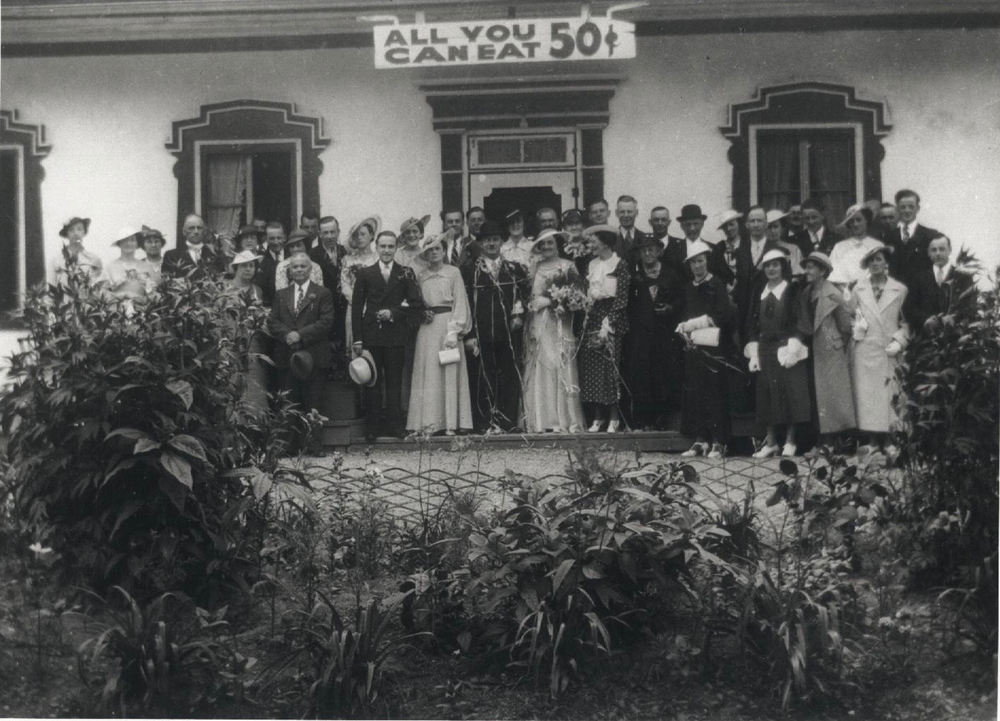 Black and white archival photo showing a bride and groom surrounded by their elegantly dressed guests in front of Auberge Baker. Everyone is looking at the camera. Behind the group, above the door, hangs a sign reading “ALL YOU CAN EAT 50₵.” In the foreground are plants and bushes.