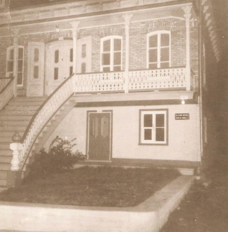 Black and white archival photo of a brick, two-storey home. The ground floor of the house, clad in white stucco, is lower than the upper storey. A staircase leads up to the veranda and to the main entrance on the upper floor. Narrow pillars support the veranda roof. A smaller door next to a window provides access to the ground floor.