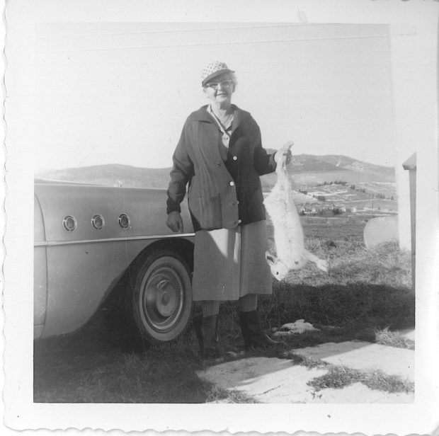 Black and white archival photo depicting an elderly Rose Lachance wearing a cap, a knit cardigan, a skirt, and rubber boots. She is holding up a dead hare in her left hand. She is standing next to a vehicle, and in the background we can see fields and hills.