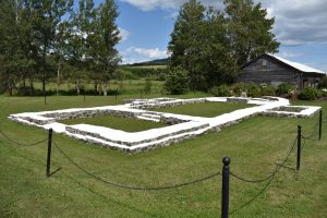 Colour photo of the stone ruins of a church on a grassy piece of land. The stones are whitewashed. From the layout of the ruins we can make out where the main entrance, nave, and chancel once were, as well as two small chapels on either side. The site, which is surrounded by trees, is cordoned off with a black, post and chain fence.