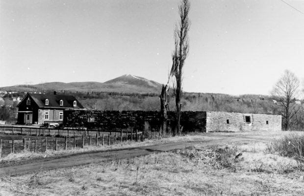 Black and white photo showing the remains of a stone building, with little more than the lower part of the walls still standing. Behind the ruins is a house, and in front stands a tree bare of leaves. In the background we can see a mountain.