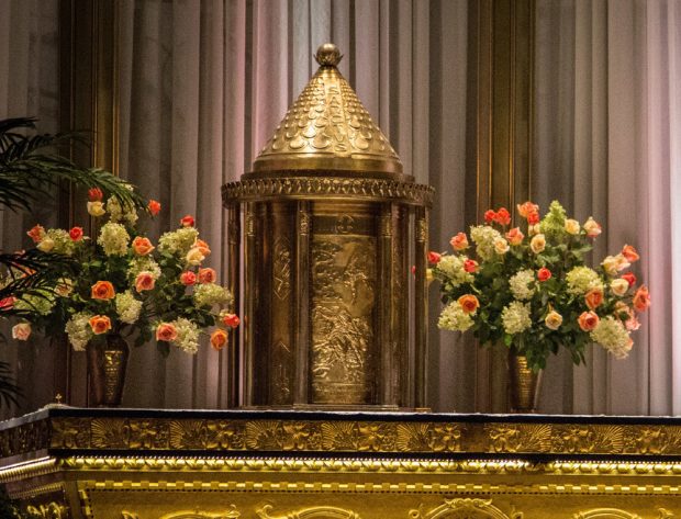 Colour photo of an ornate, embossed metal, cylindrical tabernacle set on an altar between two large vases of pink and white flowers. The top of the tabernacle is a conical shape.